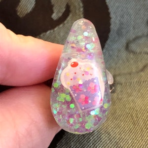Könnte beinhalten: Ein transparenter, tropfenförmiger Harz-Anhänger mit einem rosa und violetten Cupcake-Design und schimmerndem Glitzer.
