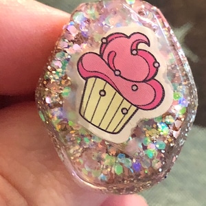 Könnte beinhalten: Ein klarer Harzanhänger mit einem rosa und gelben Cupcake-Design. Der Anhänger ist mit schillerndem Glitzer gefüllt, was einen funkelnden Effekt erzeugt. Der Anhänger wird von einer Hand gehalten. Er misst etwa 2,5 cm in der Länge.