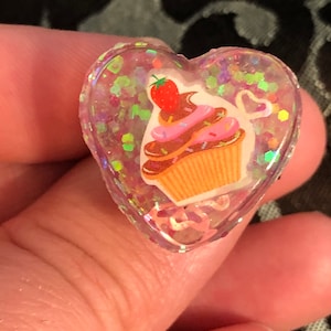 Könnte beinhalten: Ein transparenter herzförmiger Harz-Anhänger mit einem rosa und braunen Cupcake mit einer roten Erdbeere oben drauf. Der Anhänger ist mit schimmerndem Glitzer gefüllt.