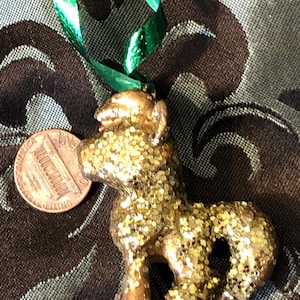 May include: A small, gold-colored horse ornament covered in glitter, with a green ribbon for hanging. A penny is next to the ornament for size comparison. The horse has a detailed design and is likely a holiday decoration.