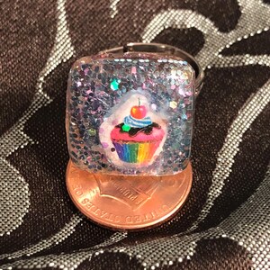 Könnte beinhalten: Ein quadratischer Ring mit einer durchsichtigen Harzplatte und einem silbernen Band. Das Harz enthält Glitzer und einen bunten Cupcake mit Regenbogen-Zuckerguss und einer roten Kirsche oben drauf.