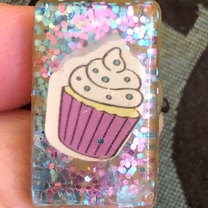 Könnte beinhalten: Ein rechteckiger, transparenter Harz-Anhänger mit einem glitzernden Hintergrund in Pink, Blau und Silber. Ein Cupcake mit weißem Frosting und lilafarbenem Zuckerguss ist in das Harz eingebettet.