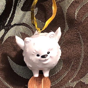 May include: White fluffy dog ornament with a gold ribbon hanging from the top. The ornament is standing on a penny.