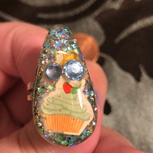 Könnte beinhalten: Ein silberner Ring mit einem klaren, tropfenförmigen Stein aus Harz. Der Stein enthält einen grünen Cupcake mit einer roten Kirsche obenauf und zwei blauen Strasssteinen. Das Harz ist mit schimmerndem Glitzer gefüllt.