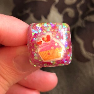 Könnte beinhalten: Ein quadratischer rosa Harz-Anhänger mit Cupcake-Design und schimmerndem Glitzer.