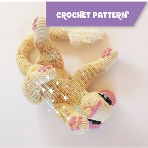 Op de afbeelding: Een gehaakt leeuwen speelgoed met een crèmekleurig lichaam en roze accenten. De leeuw ligt op zijn rug met zijn poten in de lucht. Het speelgoed heeft een pluche staart.