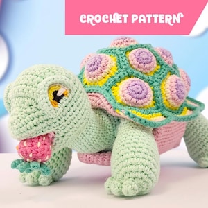 May include: A crocheted turtle toy in light green yarn, holding a small pink strawberry in its mouth. The shell features a geometric pattern with lavender, yellow, and green accents. A pink banner at the top displays the text CROCHET PATTERN.