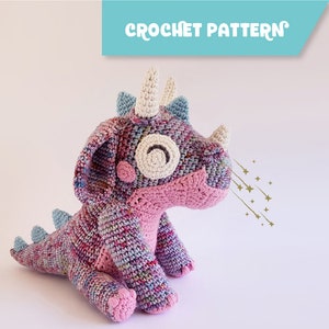 Puede incluir: Un dinosaurio de crochet rosa y azul con un cuerno blanco y ojos brillantes. El dinosaurio está sentado sobre un fondo blanco. "CROCHET PATTERN" está escrito en un cuadro azul en la parte superior de la imagen.