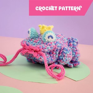 May include: A crocheted frog toy with a pink, blue and turquoise colour scheme. The frog is wearing a yellow crown and has a pink drawstring.