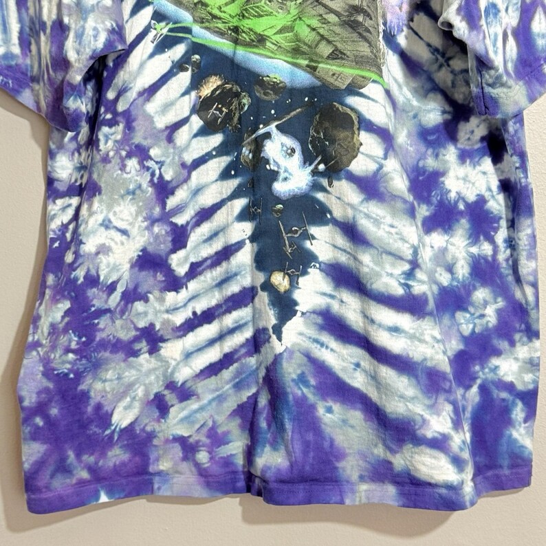 Op de afbeelding: Een tie-dye t-shirt met een ruimte-thema grafisch ontwerp. Het shirt heeft een wervelend patroon van paars, wit en blauw. De afbeelding toont astero&iuml;den, sterren en een groen en grijs landschap. Het shirt is gemaakt van een zacht, comfortabel materiaal.