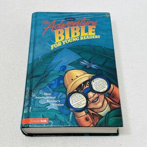 Può includere: La Bibbia dell'Avventura per i Giovani Lettori, un libro blu con un'illustrazione di un bambino che guarda attraverso un binocolo una scena di giungla. Il libro è pubblicato da Zonderkidz e presenta la versione New International Reader's.