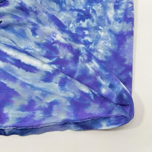 Op de afbeelding: Close-up van een tie-dye stof in tinten blauw en paars. De stof heeft een zachte, vloeiende uitstraling met zichtbare naden langs de randen. Het verfpatroon cre&euml;ert een gemarmerd effect, met lichtere en donkerdere gebieden.
