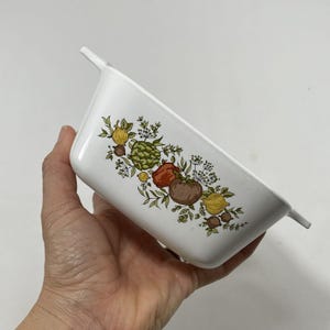 May include: White ceramic baking dish with a floral design featuring vegetables and herbs. The design includes a red pepper, a green artichoke, a yellow onion, and brown tomatoes.