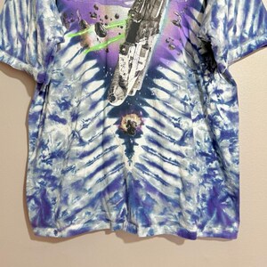 Op de afbeelding: Tie-dye T-shirt met een ruimtethema. Het shirt heeft een ruimteschipontwerp met groene laserstralen en een paars en blauw tie-dye patroon. Het shirt heeft korte mouwen.