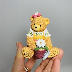 May include: A yellow teddy bear figurine wearing a green and white gingham dress and a pink flower headband. The bear is holding a small potted plant with a white flower and a miniature gardening tool.