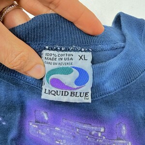Op de afbeelding: Close-up van een blauw XL Liquid Blue t-shirt label. Het label leest "100% Cotton Made in USA Care on Reverse". Het label heeft een kleurrijk ontwerp met het Liquid Blue logo.