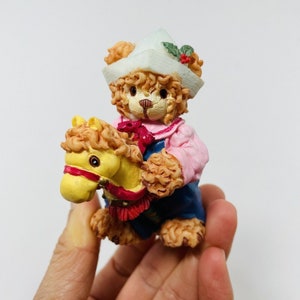 May include: A small, brown teddy bear figurine wearing a white sailor hat with a red bow and a pink shirt, riding a yellow rocking horse with a red saddle.
