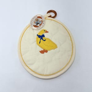 May include: Round, quilted off-white pot holder with a yellow duck embroidered on the front, wearing a blue bow. The pot holder has a yellow border and a brown loop for hanging. A tag with text is attached.