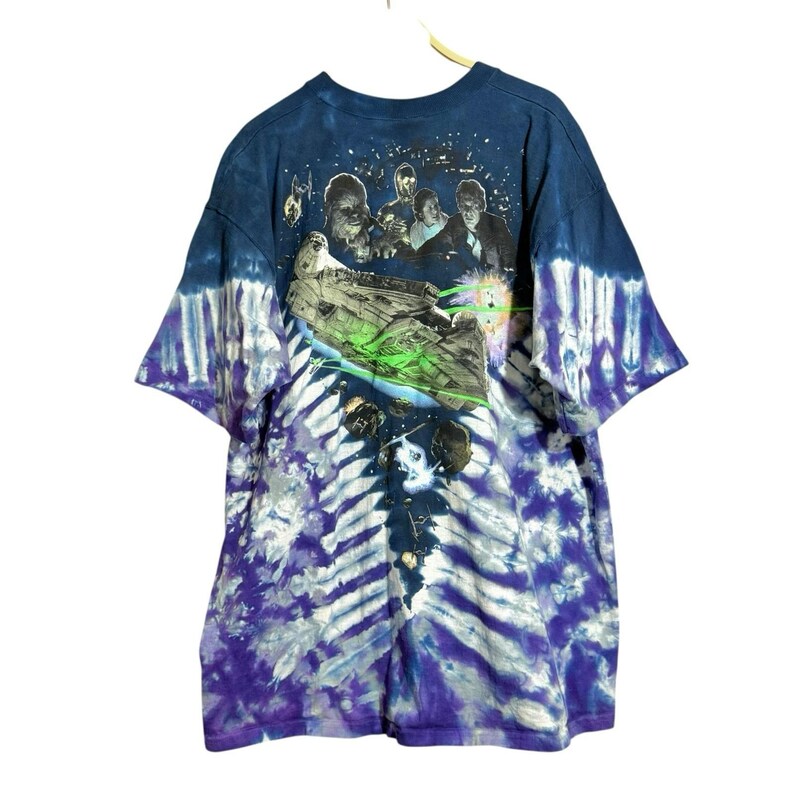 Op de afbeelding: Tie-dye T-shirt met een Star Wars-afbeelding. Het ontwerp toont de Millennium Falcon die lasers afvuurt, met personages als Chewbacca, Han Solo en Luke Skywalker. Het shirt heeft een blauw en paars tie-dye patroon.