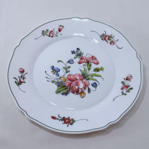 May include: White ceramic plate with a scalloped edge and a green trim. The plate features a central floral design with pink, red, blue, and yellow flowers. Additional floral accents are scattered around the rim.