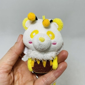 May include: A white plush toy shaped like a bee with yellow accents and a brown honeycomb pattern. The toy has a cute, cartoon-like face with pink cheeks and yellow eyes.