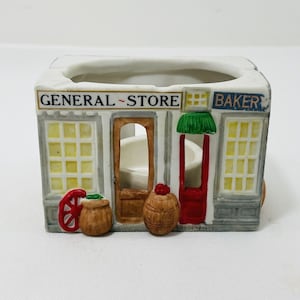 May include: A white ceramic candle holder shaped like a general store and bakery. The front of the building has two windows, a door, and a sign that reads "GENERAL STORE" and "BAKER". There are two barrels and a wheel in front of the building.