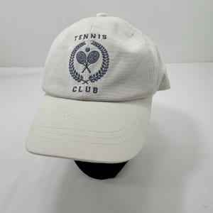 May include: Cream-coloured baseball cap with a curved peak. The cap has the words "TENNIS CLUB" embroidered in navy blue, along with a tennis racket and ball emblem. The hat is made of a soft, textured fabric.