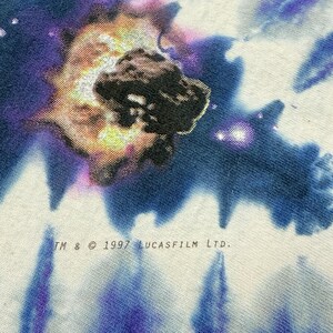 Op de afbeelding: Close-up van een wit t-shirt met een kleurrijk grafisch ontwerp. Het ontwerp bevat een donkere, getextureerde vorm omgeven door een levendige explosie van kleuren, met paarse en blauwe tinten. De tekst "TM & &copy; 1997 LUCASFILM LTD." staat onderaan.