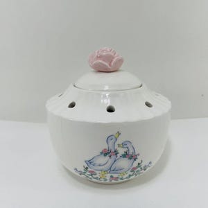May include: White ceramic pot with a pink rose lid and a floral design featuring two ducks. The pot has holes in the bottom and sides.
