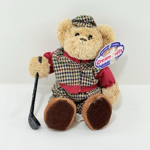 May include: A soft toy teddy bear wearing a golf outfit, including a houndstooth vest and cap, holding a golf club. The bear has brown fur and wears a red shirt. A tag reads "Cream Puffs".