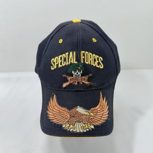Peut inclure: Casquette de baseball bleu marine avec l'inscription "SPECIAL FORCES" brodée en or. La casquette présente un aigle brodé aux ailes déployées et un crâne portant un béret et des fusils croisés.