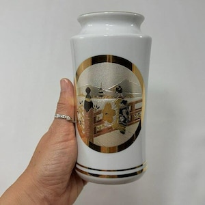 Puede incluir: Jarrón de cerámica blanca con borde dorado y diseño circular decorativo dorado y plateado. El diseño presenta dos figuras con vestimenta tradicional japonesa en un puente, con una pagoda al fondo.