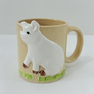 May include: Tan ceramic mug with a white pig figurine on the front. The pig has brown hooves and a black eye. Green grass decorates the bottom of the mug. The mug has a curved handle.