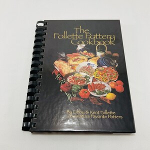 Könnte beinhalten: Ein schwarzes Kochbuch mit goldenem Titel "The Follette Pottery Cookbook", das ein farbenfrohes Bild eines Essens auf einem Holztisch zeigt. Der Text "By Libby & Kent Follette Louisiana's Favorite Potters" ist unter dem Titel gedruckt.