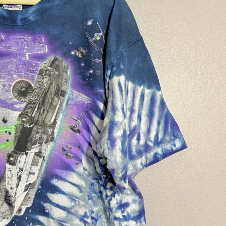 Op de afbeelding: Blauw tie-dye T-shirt met een ruimtesc&egrave;ne, inclusief een groot ruimteschip, straaljagers en een paarse en blauwe galaxy-achtergrond. Het shirt heeft een ronde hals en korte mouwen.