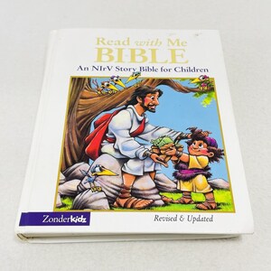 May include: A white book with a colorful illustration of Jesus giving a basket of food to a child. The book's title is "Read with Me BIBLE" and the subtitle is "An NIrV Story Bible for Children". The book is published by ZonderKidz and is revised and updated.