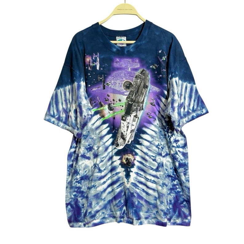 Op de afbeelding: Tie-dye T-shirt met een Star Wars-afbeelding. Het ontwerp toont een ruimtegevechtsc&egrave;ne met een groot ruimteschip, laserstralen en andere ruimtevaartuigen tegen een paarse en blauwe achtergrond. Het shirt heeft een ronde hals en korte mouwen.