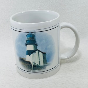Könnte beinhalten: Weiße Keramik-Kaffeetasse mit einem Leuchtturm-Motiv. Der Leuchtturm ist weiß mit schwarzen Streifen und einem blauen Dach. Das Motiv ist von einem schwarzen Rahmen umgeben.