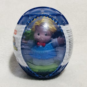 May include: A Fisher-Price Little People toy in a clear plastic egg-shaped container. The toy figure has blonde hair, a red bow tie, and a blue outfit. The container is blue and green.
