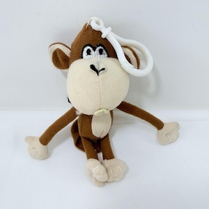 May include: A plush monkey keychain with brown fur, tan face, ears, hands, and feet. The monkey has embroidered eyes and a white plastic clip for attaching to keys or a bag. The monkey's arms and legs are outstretched.