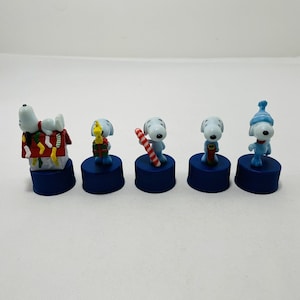 以下が含まれることがあります： 青いボトルキャップの上に、青と白のピーナッツのキャラクターのフィギュアが5体あります。キャラクターはホリデーの服装をして、ホリデーのアイテムを持っています。