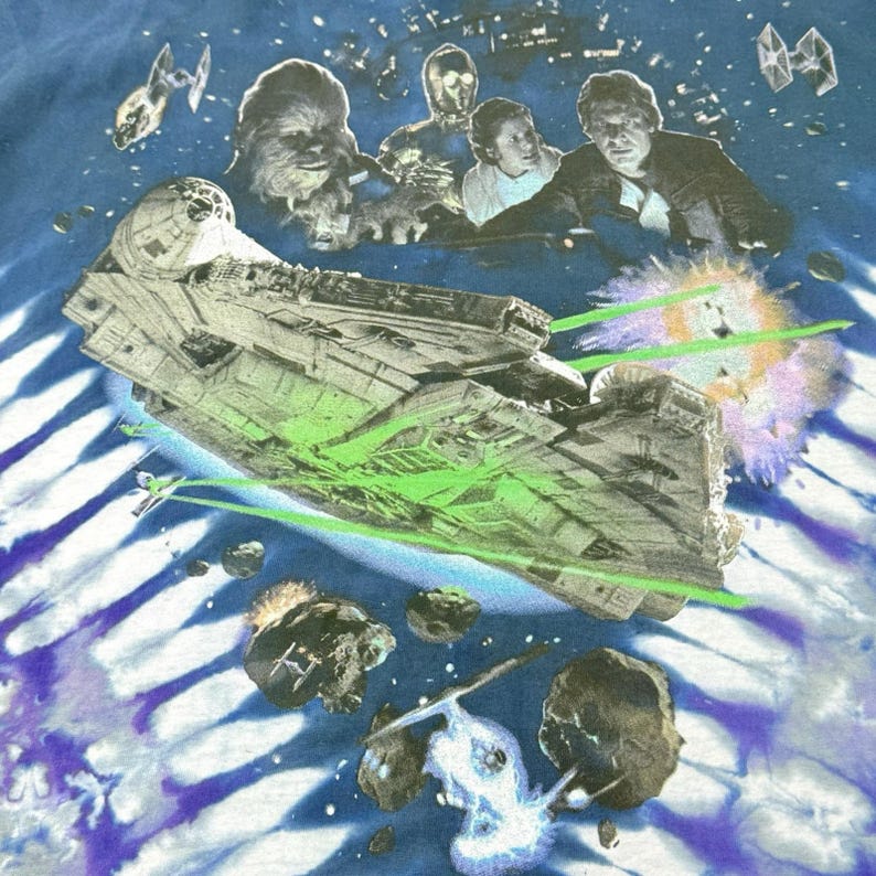 Op de afbeelding: Tie-dye T-shirt met een Star Wars-ontwerp. Het ontwerp bevat de Millennium Falcon, Chewbacca, C-3PO en Han Solo. Het shirt heeft een blauw-witte tie-dye achtergrond met ruimte-elementen en groene laserstralen.