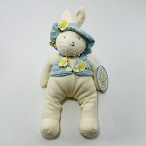 Puede incluir: Un conejito de peluche blanco con un sombrero a rayas azul y blanco con flores amarillas y un chaleco azul con flores amarillas. El conejito tiene una campana en su sombrero y una etiqueta que dice "Plush Toys by The Yard" y "Buttercup".