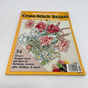 Puede incluir: Una revista vintage "Cross-Stitch Bazaar" con un diseño floral de punto de cruz en la portada. La portada es amarilla y tiene el texto "McCall's Creative Crafts" y "¡34 diseños nuevos!" Se ve un manojo de hilos de bordar coloridos.