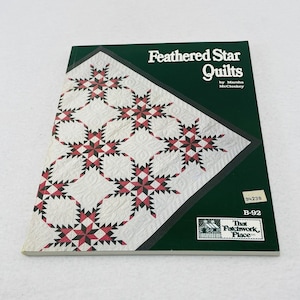 Puede incluir: Un libro titulado "Feathered Star Quilts" de Marsha McCloskey. La portada presenta un diseño de edredón con patrones de estrellas rojas, negras y blancas sobre un fondo blanco. El libro tiene una cubierta verde oscuro con el título en blanco.