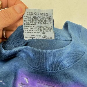 Op de afbeelding: Close-up van een blauwe t-shirtkraag met een wasvoorschriftenlabel. Het label geeft wasinstructies, waaronder apart wassen en het gebruik van chloorvrije bleekmiddelen. Op het label staat ook dat het kledingstuk in de VS is gemaakt.