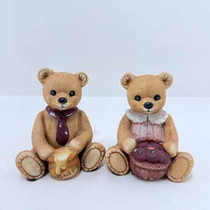Könnte beinhalten: Zwei Keramik-Teddybär-Figuren. Ein Bär trägt eine bordeauxfarbene Krawatte und hält einen Honigtopf mit dem Wort "HONEY" darauf. Der andere Bär trägt ein rosa Kleid und hält einen Korb mit lila Früchten. Beide Bären sitzen.