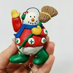 May include: A snowman ornament with a red and white sweater, a blue scarf, and a green broom. The snowman has a white face with black eyes and a carrot nose. The ornament is decorated with plus signs.