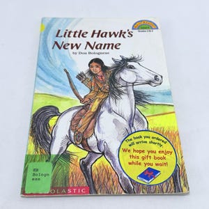 Puede incluir: Un libro infantil titulado "Little Hawk's New Name" de Don Bolognese. La portada presenta una ilustración de un niño nativo americano a caballo con arco y flechas. El libro tiene una pegatina amarilla y azul que dice "El libro que pediste llegará pronto."