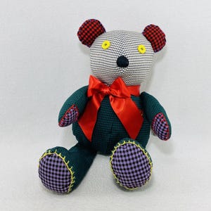 May include: A handmade teddy bear with a black and white checkered head, red and black plaid ears, and a black nose. The bear has a green plaid body, red bow, and purple and black checkered feet. Yellow stitching and button eyes add detail.